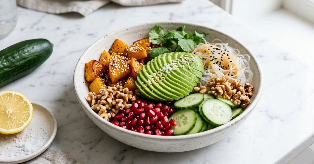 Bowl filled with roasted squash pieces sprinkled with sesame seeds, half an avocado, sliced cucumbers, pomegranate seeds, walnuts, and glass noodles topped with black sesame seeds.